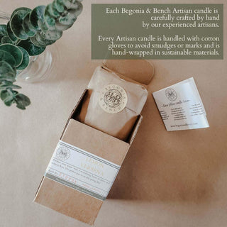 A box with a partially opened Desert Spice™ artisan candle from Begonia & Bench® sits on a wooden surface. Surrounding the candle are a eucalyptus branch and hints of amber. The image text emphasizes the candles' handcrafted nature and sustainable packaging.