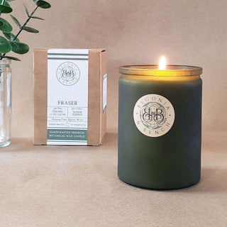 A lit Yuzami™ 12oz finely crafted scented candle by Begonia & Bench® in a dark green jar sits on a beige surface. Behind it, there's a cardboard box with the same branding indicating the candle is a "Fraser" scent featuring notes of fir, cedar, grand fir, and green apple. A sprig of leaves rests in a glass jar on the left.