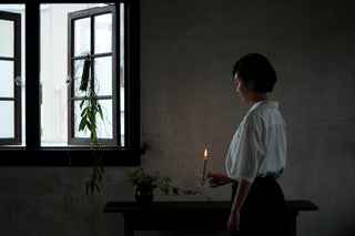 A person stands indoors, holding a lit candle. They are near a table with a small plant, and they are looking at the candle flame. The room is dimly lit, with light coming through partially open windows. A vine with leaves hangs near the window. The person is dressed in a white shirt and dark pants.