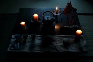 A serene tea setup on a dark wooden table illuminated by five candles. Central to the arrangement is a traditional black teapot. Surrounding it are cups, a small bowl, and a cloth. On the right side, a dried plant hangs next to a candle, adding to the tranquil ambiance.