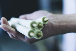 Close-up of a person's hand holding four rolled up white candles with visible wicks. The candles are arranged in a bundle and appear to be new and unused. The background is blurred, drawing focus to the hand and candles. The lighting is soft, with a cool, muted tone.