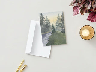 A Greeting Card showcasing a serene watercolor painting of a forest landscape with a river is lying next to a plain white envelope. Nearby, there's a lit scented candle in a glass holder, two yellow-tipped pens, and some red-and-green leaves, all arranged on a light-colored surface.