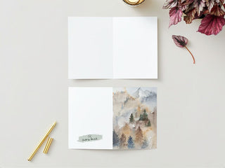 A flat-lay image of the Greeting Card set on a light surface. The card is open at the top with a blank interior, and its front cover displays a watercolor painting of a misty forest with trees and mountains. Surrounding it are gold pens, red leaves, and a fall candle that add warmth to the scene.