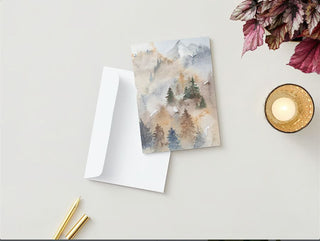 The Greeting Card, featuring an illustration of a misty, forested mountain scene, is displayed on a white surface next to an envelope, a gold pen without a cap, and another pen with its cap on. To the right of the card sits a lit fall candle in a textured glass holder and some variegated leaves.