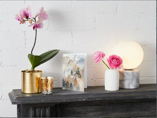 A mantelpiece displays a gold pot with a pink orchid, two gold candle holders, a framed nature painting of mountains, a white vase with pink ranunculus flowers, and a marble-based globe lamp. A Greeting Card would perfectly complement the elegant scene set against the white brick background.