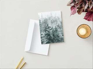 A Greeting Card featuring a watercolor painting of a misty evergreen forest is placed on a white surface next to an open white envelope. Nearby, two gold pens, a lit fall candle in a decorative holder, and a bunch of purple and green leaves complete the picturesque scene.