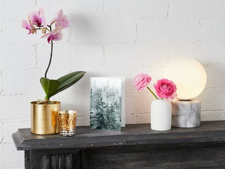 A decorative setup on a mantel showcases a gold pot with a blooming pink orchid, a silver candle holder with a scented candle, the Greeting Card featuring a forest design, and a white vase holding pink flowers. A marble-based lamp with a glowing round bulb completes the arrangement against a white brick wall.