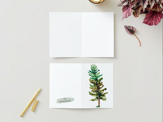 The Greeting Card lies open on a light grey surface, showcasing an upright pine tree illustrated in watercolor on the front. The inside of the card is blank. Surrounding it are a gold pen, another gold object, some red and green leaves, and a scented candle to create a cozy atmosphere.