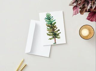 A Greeting Card showcasing a watercolor painting of a green pine tree is displayed partially inside a white envelope. Next to it, there is a lit, scented candle in a glass holder with a textured surface, a gold pen, and an arrangement of red and green leaves on a light beige surface.