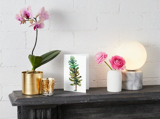 A decorative scene on a black mantelpiece features a golden pot holding an orchid with light purple flowers, a glass candle holder, Greeting Card with a watercolor tree design, a white vase with pink flowers, and a marble lamp with a glowing round bulb, set against a white brick wall.