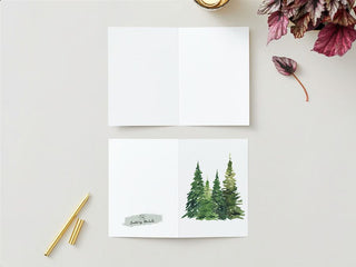 A blank Greeting Card is open on a light surface with a painting of three pine trees on the right page. Another identical closed card is beneath it, displaying the back cover which features the same tree design and the text "Soulful by Camila" above a brush stroke. Nearby, there is a pen, some plant leaves, and a lit fall candle adding warmth to the scene.