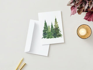 A Greeting Card from Michelle's Greeting Cards depicting three watercolor evergreen trees lies on top of a matching white envelope. Beside them, there is a golden pen and a matching pencil. To the right, a lit candle in a gold holder and a cluster of purple and green leaves can be seen on a light beige surface.