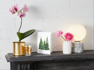  A white brick wall backdrop features a black mantel adorned with decorative items: a potted orchid with purple flowers in a gold planter, two small gold glass votives, our charming Greeting Card showcasing painted evergreen trees, pink flowers in a white vase, and a marble lamp with a glowing orb. A fall candle adds warmth to the cozy scene.