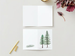 A minimalistic Greeting Card set on a light surface. The top card is open, revealing a blank interior. The bottom card, from Michelle's Greeting Cards, features a watercolor illustration of two evergreen trees and text that appears to read "Best wishes" or similar. Nearby are a gold pen, a golden paper clip, and some red-tinted leaves.