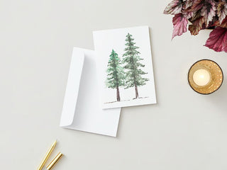 A Greeting Card adorned with a watercolor painting of two evergreen trees is displayed beside a white envelope. On the light gray surface around them, you'll find two gold pens and a lit, scented candle in a textured glass holder. A few reddish autumn leaves are visible in the top right corner.