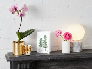 A mantel adorned with a potted orchid in a gold container, gold votive candle holders, a Greeting Card featuring two pine trees, and a vase with pink flowers. A marble base lamp with a round white light sits on the right end against a white brick wall background.