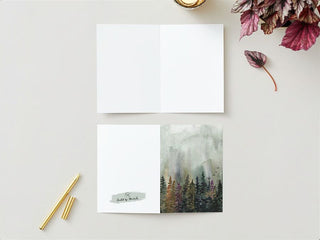 A Greeting Card lies open on a gray surface, revealing its white blank inner pages. Adjacent to it is the cover of the Greeting Card, which features a watercolor forest scene with green trees and a misty background. Nearby, there is a gold pen with a matching cap, some red-tinted leaves, and a cozy fall candle adding warmth to the scene.