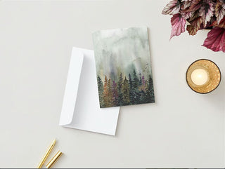 A Greeting Card adorned with a watercolor painting of a forest rests on a white envelope on a light gray surface. To the left, there is a gold pen and a pencil. In the top right corner, purple leaves are scattered, while in the bottom right corner, a lit fall candle in a golden holder adds warmth to the scene.