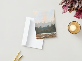 A Greeting Card depicting a tranquil forest and mountain landscape painting rests partially inside a white envelope on a light beige surface. Next to it, there are two gold pens, a lit fall candle in a glass holder, and in the upper right corner, an arrangement of purple and green leaves.