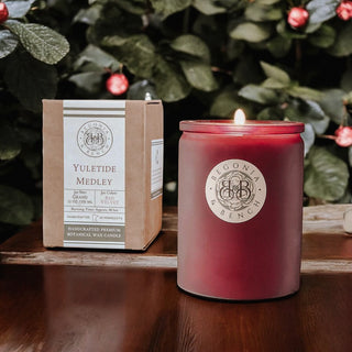 The Yuletide Medley™ scented candle by Begonia & Bench® rests on a wooden surface, casting a warm glow in its frosted glass jar and filling the air with holiday fragrances. The label displays "Begonia & Bench," while the accompanying box details the festive scent of grand fir, cider, and velvet. In the background, green foliage adorned with small red flowers adds to the seasonal ambiance.