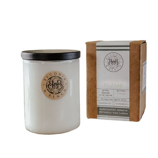 A 12oz Yuzami™ scented candle by Begonia & Bench® in a white jar with a black lid is placed beside a brown box, reflecting minimalist design. The candle displays the "Begonia & Bench" branding within its round logo, while the box features "Yuzami," highlighting the phrase "Handcrafted Premium Botanical Wax Candle"—perfect for connoisseurs of exquisite artisan candles.