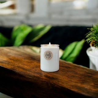 A Yuzami™ - 12oz Finely Crafted Scented Candle by Begonia & Bench® sits elegantly in a glass holder on a rustic wooden surface. The candle's label prominently displays the Yuzami logo. In the softly blurred background, green leaves and a white planter contribute to the natural aesthetic of the scene. Warm, gentle lighting evokes a calming ambiance, reminiscent of soothing matcha moments.