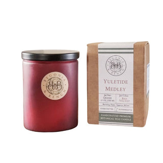 A red candle in a glass jar with a black lid bears the Begonia & Bench® label. Next to it, a brown box with "Yuletide Medley™" reveals information about this holiday fragrance. Weighing 12 oz and offering a 75-hour burn time, this handcrafted artisan candle is made from premium botanical wax sourced from Minnesota.