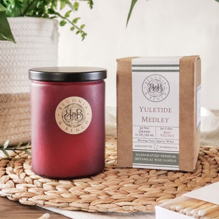 A decorative red candle jar rests on a woven mat next to its box, which is labeled "Yuletide Medley™ - 12oz Finely Crafted Scented Candle By Begonia & Bench®." This artisan candle boasts festive fragrances and includes information about its scent and burn time. In the background, a green plant and wooden beads enhance the cozy atmosphere.