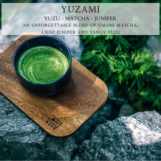 A finely crafted 12oz Yuzami™ scented candle by Begonia & Bench® rests on a wooden tray, reminiscent of a blue bowl filled with vibrant green matcha. Set against a stone backdrop and lush greenery, the scene features the phrase: "YUZAMI: A unique fusion of umami matcha, crisp juniper, and tangy yuzu.