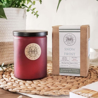 A burgundy candle featuring a golden emblem sits next to a cardboard box labeled with "Snow Frost™ - 12oz Finely Crafted Scented Candle By Begonia & Bench®." This artisan candle, handcrafted from botanical wax, is housed in a red velvet jar and infused with the Snow Frost scent. A woven mat and greenery serve as a calming backdrop.