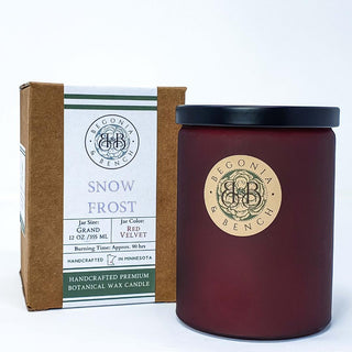 A red candle jar marked "Begonia & Bench" is placed next to a brown box featuring "Snow Frost™ - 12oz Finely Crafted Scented Candle By Begonia & Bench®" along with "Handcrafted Premium Artisan Candle." Both items boast details such as jar size, color, and a 90-hour burn time, showcasing a circular ornate emblem that exudes Red Velvet luxury.